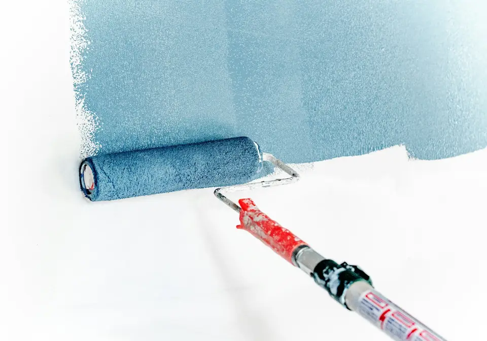 Painter rolling a fresh coat on a living room wall with furniture covered and trim taped off